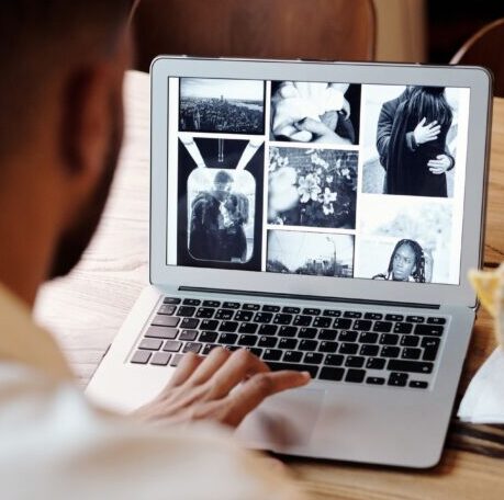 Person holding a sandwich while browsing images on a laptop at a wooden table.